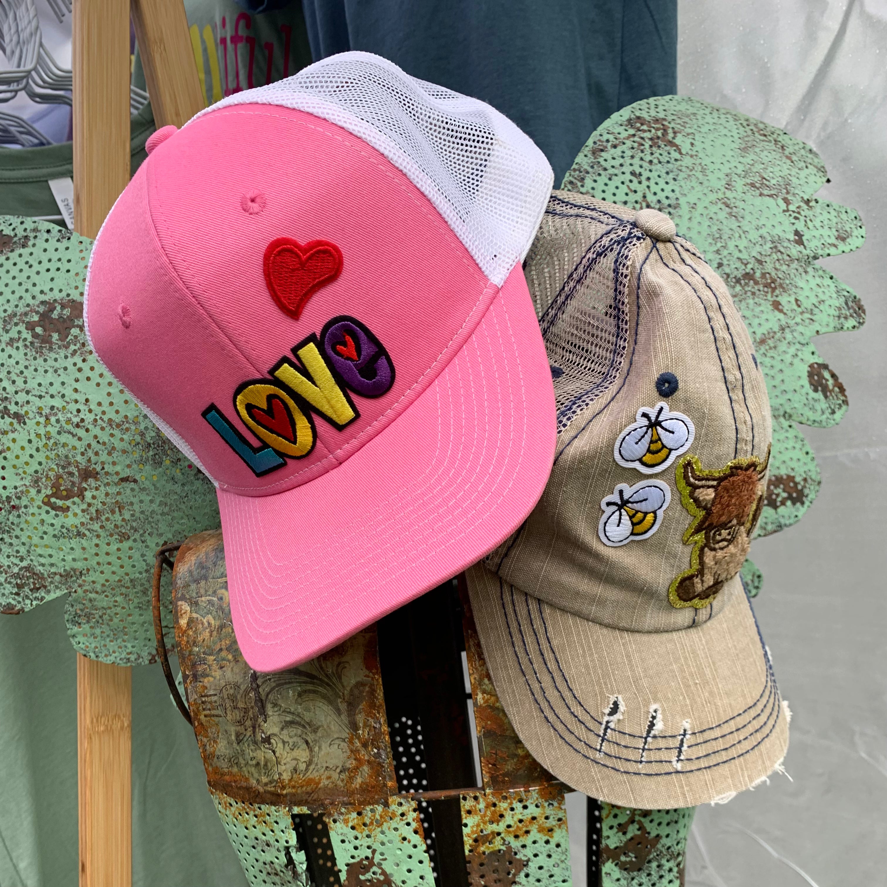 Pink cap with colorful 'LOVE' text and red heart, beige cap with embroidered designs on a stand.
