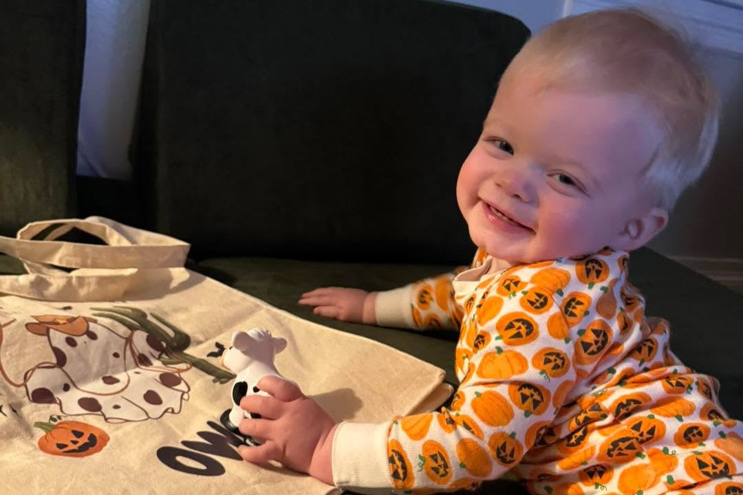 Baby in Halloween-themed outfit sitting next to a tote bag with pumpkins on a couch.