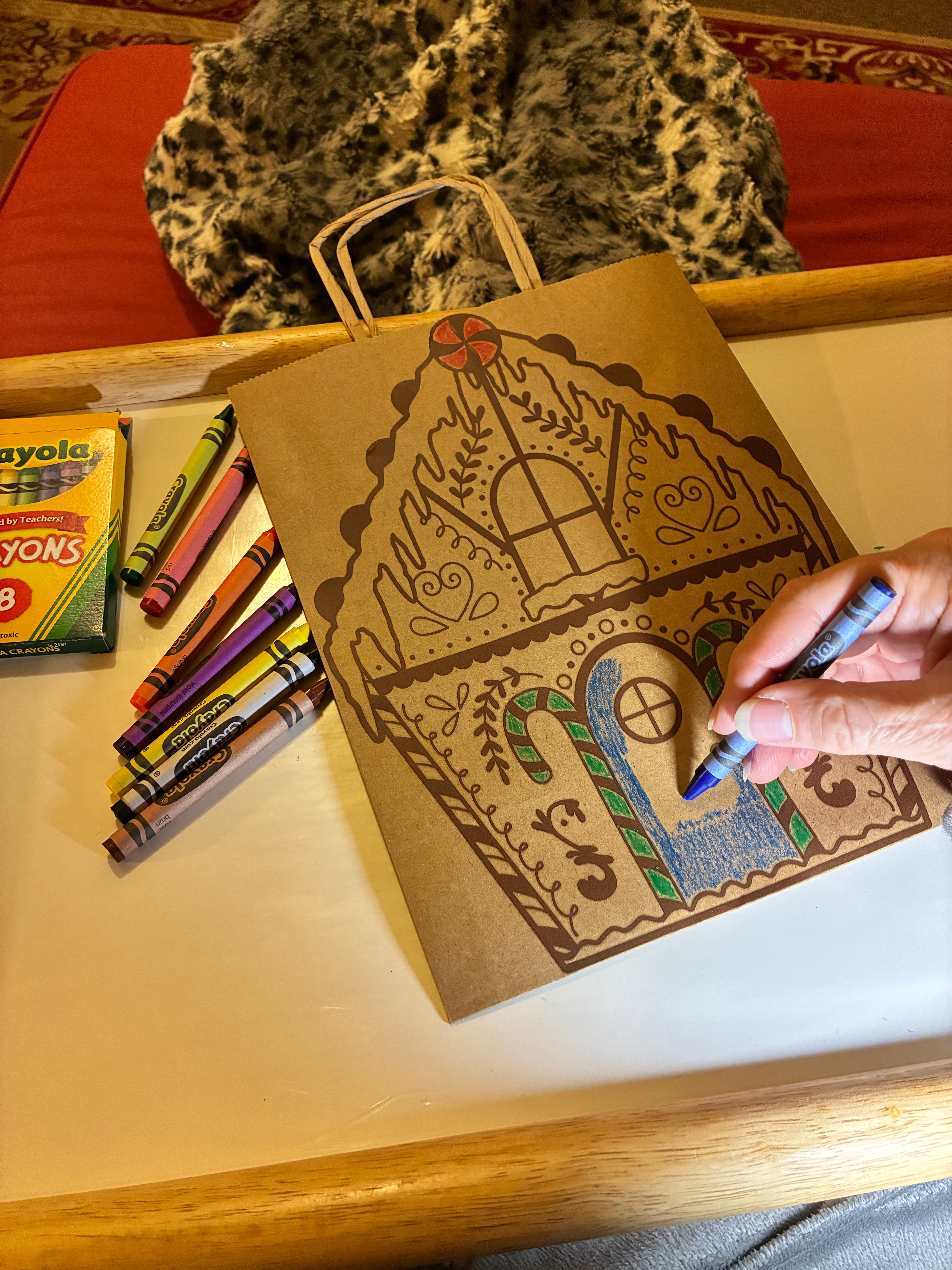Person coloring a goningerbread house with crayons on a table.