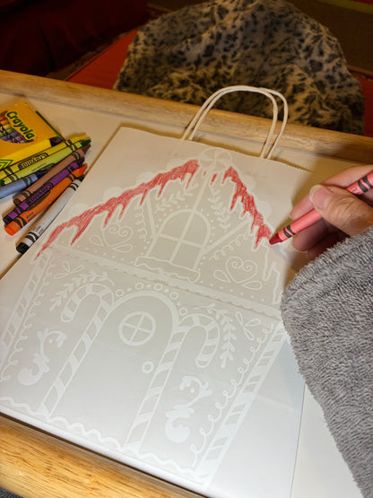 Person coloring on a gingerbread house with Crayola crayons on a wooden table.